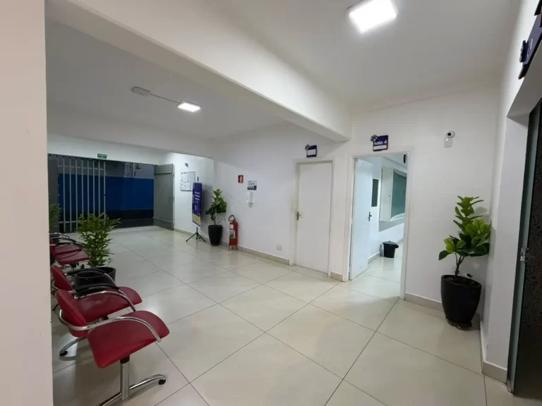 Corredor interno com cadeiras de espera vermelhas, plantas e as portas das salas