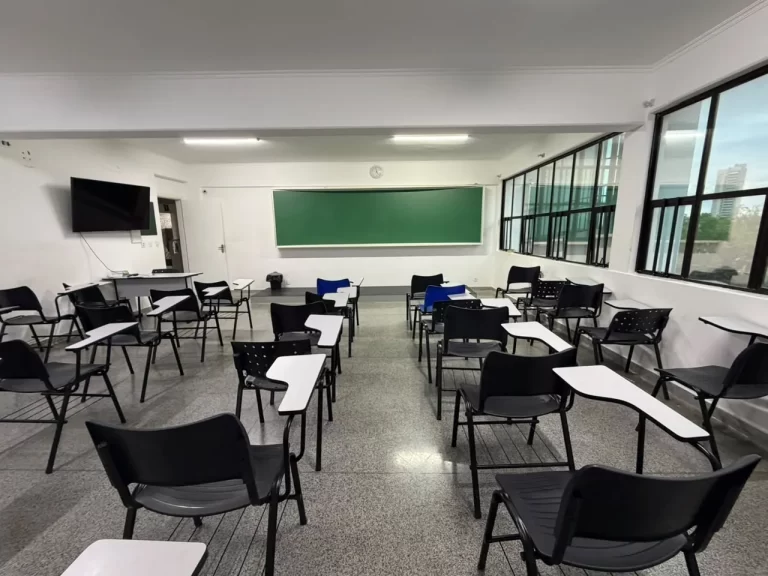 Sala de aula ampla com janelas laterais, lousa verde, televisão e carteiras universitárias