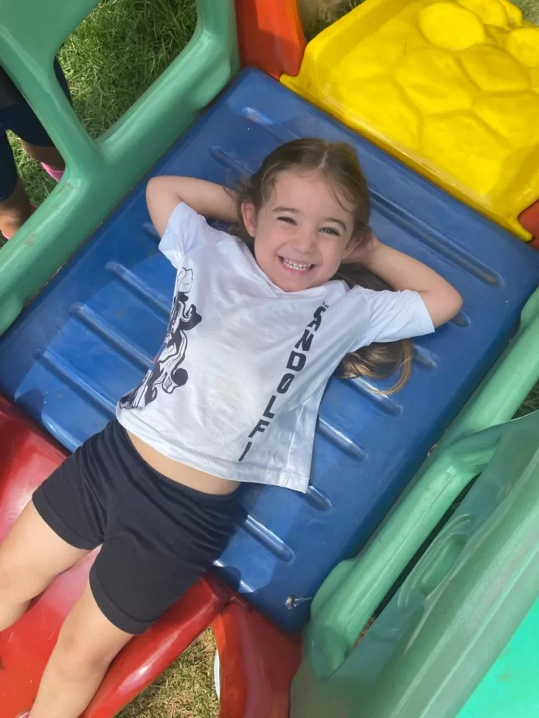 Aluna da Educação Infantil com a camiseta do Gandolfi, deitada e sorrindo no escorregador do parquinho